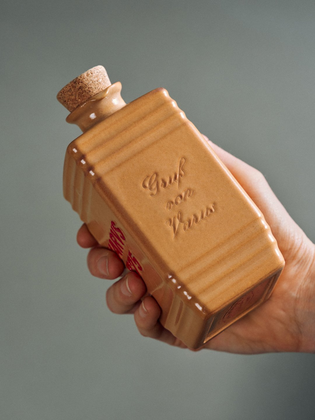 Hand holding tan rectangular handled bottle against muted gray background, Varus 775 by Olssøn Barbieri, Lübbecke, Olssøn Barbieri / Varus Brennerei, Lübbecke, Germany