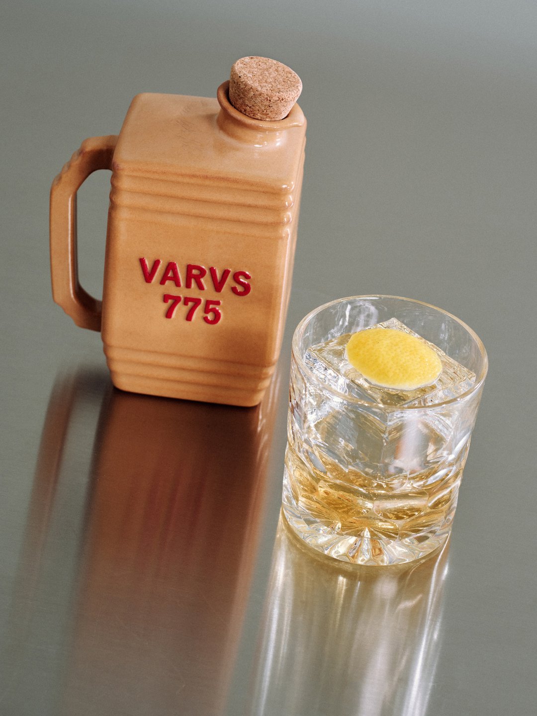Tan handled bottle beside iced drink with citrus on reflective surface, Varus 775 by Olssøn Barbieri, Lübbecke, Olssøn Barbieri / Varus Brennerei, Lübbecke, Germany
